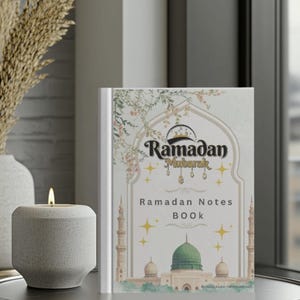 Könnte beinhalten: Ein Ramadan-Notizbuch mit dekorativem Einband. Der Einband zeigt die Worte "Ramadan Mubarak" und Illustrationen einer Moschee, Sterne und florale Elemente. Eine brennende Kerze und eine Vase befinden sich im Hintergrund.