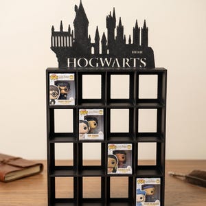 Peut inclure: Étagère noire à 12 compartiments, surmontée d'une silhouette noire du château de Poudlard et du mot "HOGWARTS". L'étagère contient plusieurs figurines Harry Potter Funko Pop dans leurs boîtes d'origine. L'étagère est en bois.