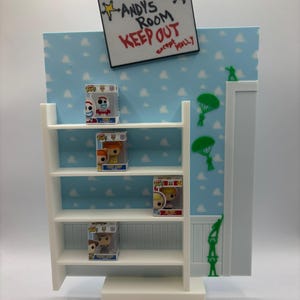 Peut inclure: Étagère blanche à quatre niveaux, exposant des figurines Funko Pop. L'arrière-plan présente un ciel bleu avec des nuages blancs et une pancarte indiquant "ANDY'S ROOM KEEP OUT except MOLLY". Des soldats et parachutes verts sont également visibles.