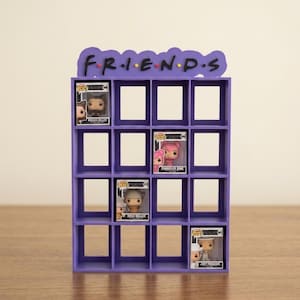 Peut inclure: Une étagère d'exposition violette avec le mot "FRIENDS" en haut. L'étagère comporte plusieurs compartiments carrés, dont certains contiennent des figurines Funko Pop. Les figurines représentent divers personnages de la série télévisée "Friends".