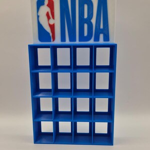 Peut inclure: Un logo NBA bleu et blanc avec une silhouette de joueur de basket-ball est affiché au-dessus d'une étagère bleue à plusieurs compartiments. L'étagère comporte 16 ouvertures carrées, parfaites pour exposer de petits objets. Le design est un hommage à la NBA.