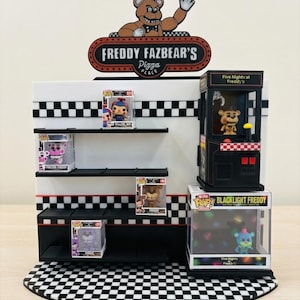 Peut inclure: Une présentation avec un panneau Freddy Fazbear's Pizza Place et plusieurs figurines Funko Pop de Five Nights at Freddy's. L'étalage comprend une machine à griffes, des étagères et un sol à carreaux. Le panneau présente un ours brun.