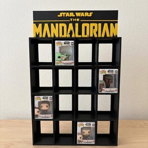Peut inclure: Étagère noire pour figurines Funko Pop, avec le logo Star Wars: The Mandalorian. L'étagère contient plusieurs figurines, dont Grogu, Boba Fett et Fennel Shand. L'étagère est en matériau noir et est posée sur une surface claire.