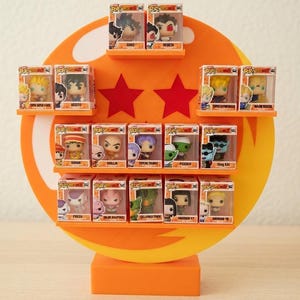 Peut inclure: Un présentoir orange avec plusieurs étagères, contenant diverses figurines Funko Pop dans des boîtes transparentes. Le présentoir présente un grand cercle orange avec deux étoiles rouges et une base rectangulaire. Les figurines sont de la série Dragon Ball Z.
