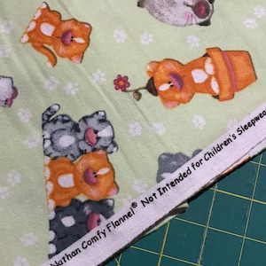 May include: A light green flannel fabric featuring a repeating pattern of cartoon cats, paw prints, and fish bones. The white selvage edge displays the text: A.E. Nathan Comfy Flannel Not Intended for Children's Sleepwear.