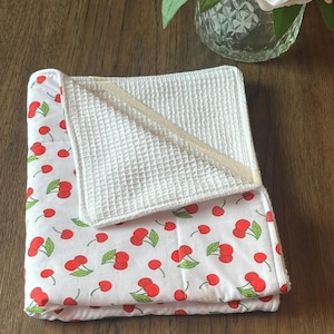 May include: A folded white baby blanket with a red cherry pattern. The blanket has a white waffle-weave section and a cream-colored trim. The blanket is folded on a wooden surface.
