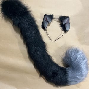 May include: A black and gray faux fur cat tail and matching ear headband set. The tail is long and fluffy, transitioning from black to gray. The headband features two small, gray and black cat ears.