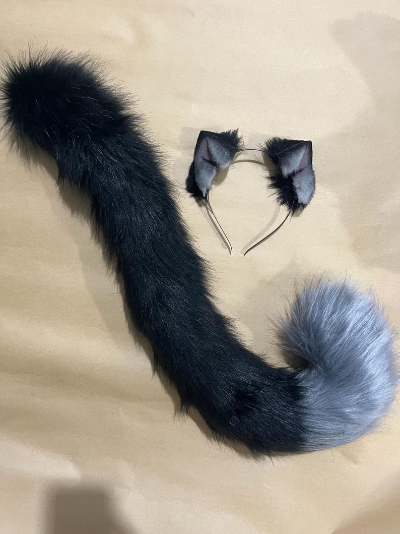 May include: A black and gray faux fur cat tail and matching ear headband set. The tail is long and fluffy, transitioning from black to gray. The headband features two small, gray and black cat ears.