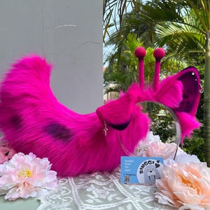 May include: A bright hot pink faux fur headband featuring two tall antennae with round tips and large, pointed ears. The ears are accented with dark purple tips, silver star-shaped charms, and a small hoop earring. A blue business card reads Binggi.