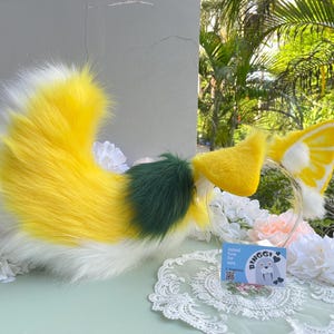May include: A faux fur animal tail and matching headband with ears. The tail features a gradient of white, bright yellow, and a dark green patch. The headband has yellow and white ears with a citrus slice pattern and a small blue tag reading Binggi.