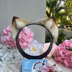 May include: A black headband with attached faux fur animal ears. The ears are tan and white. The headband is displayed with pink and white flowers and a business card that says "Animal faux fur ears".