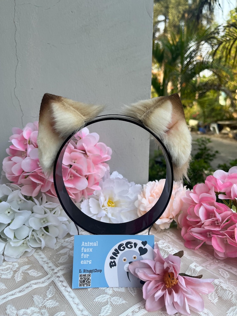 May include: A black headband with attached faux fur animal ears. The ears are tan and white. The headband is displayed with pink and white flowers and a business card that says "Animal faux fur ears".