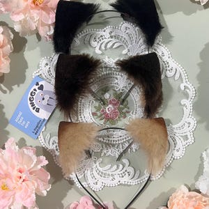 May include: Three faux fur animal ear headbands in black, brown, and tan. The ears are attached to thin black headbands. A small blue card with a cartoon animal and text is visible. The headbands are displayed on a white lace doily, surrounded by pink flowers.