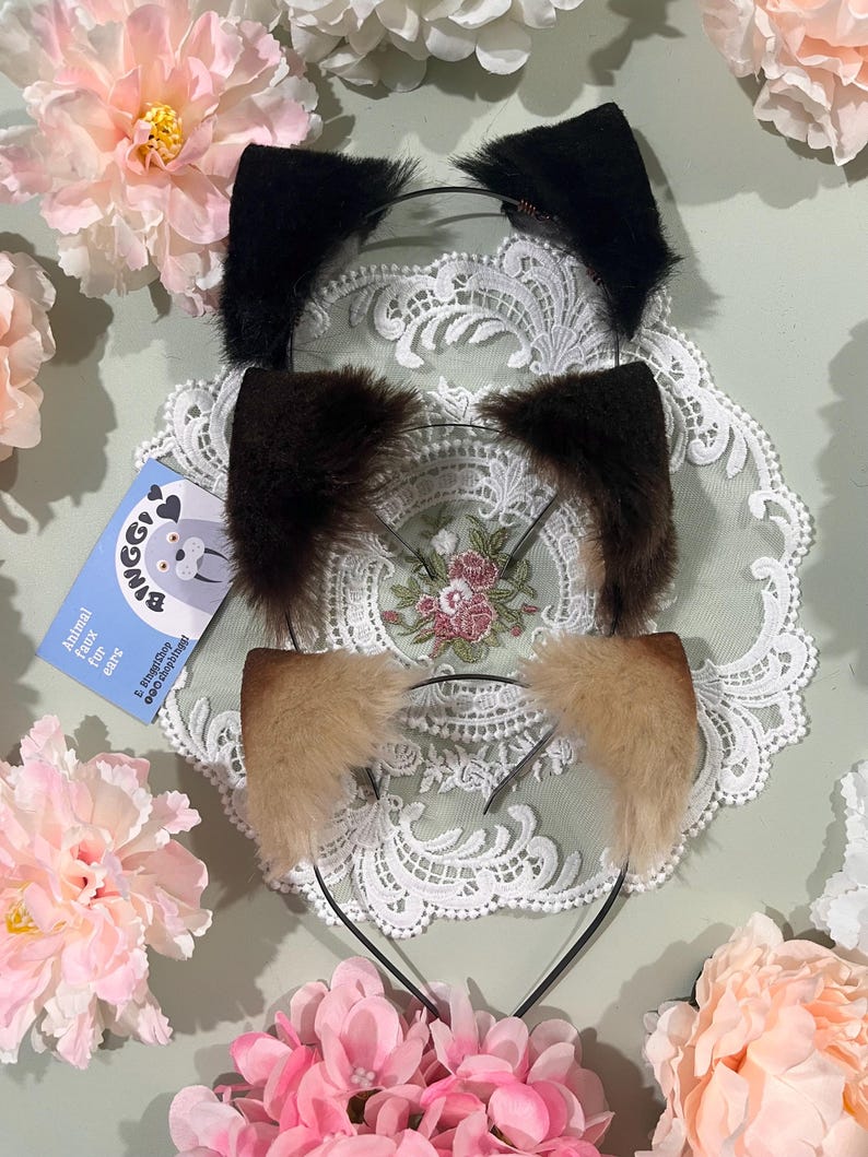 May include: Three faux fur animal ear headbands in black, brown, and tan. The ears are attached to thin black headbands. A small blue card with a cartoon animal and text is visible. The headbands are displayed on a white lace doily, surrounded by pink flowers.