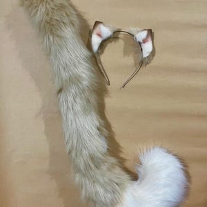 May include: A cosplay set featuring a fluffy, light brown and white cat tail and matching headband with ears. The tail is long and tapered, with a white tip. The ears are light brown with pink inner details, attached to a thin metal headband.