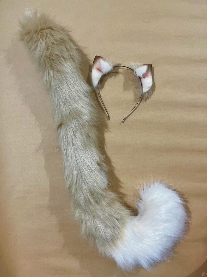 May include: A cosplay set featuring a fluffy, light brown and white cat tail and matching headband with ears. The tail is long and tapered, with a white tip. The ears are light brown with pink inner details, attached to a thin metal headband.