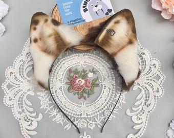 May include: A headband featuring two faux fur animal ears with a brown and white spotted pattern. The ears are attached to a thin black metal band. A small silver-toned paw print charm hangs from the right ear. A blue card with the text Binggi is visible.