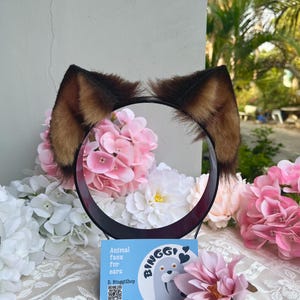 May include: A black headband with brown and tan faux fur animal ears. The ears are surrounded by pink and white flowers. A blue card with the text "Animal faux fur ears" and a walrus illustration is in front of the headband.