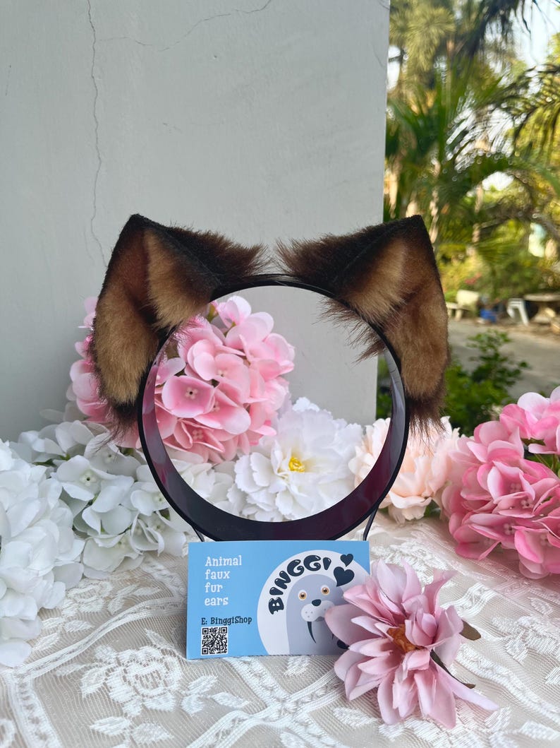 May include: A black headband with brown and tan faux fur animal ears. The ears are surrounded by pink and white flowers. A blue card with the text "Animal faux fur ears" and a walrus illustration is in front of the headband.
