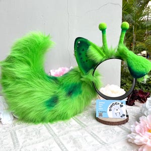Puede incluir: Una cola de piel sintética verde brillante y una diadema a juego con dos antenas altas y orejas puntiagudas. Las orejas tienen manchas verde oscuro, pequeños dijes de estrellas plateadas y cruces colgantes. Una tarjeta muestra el texto Binggi.