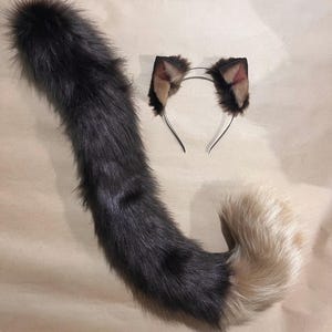 May include: A faux fur cat tail and matching headband with cat ears. The tail is primarily dark gray, transitioning to a light tan at the tip. The ears are dark brown with tan and pink accents, attached to a silver headband.