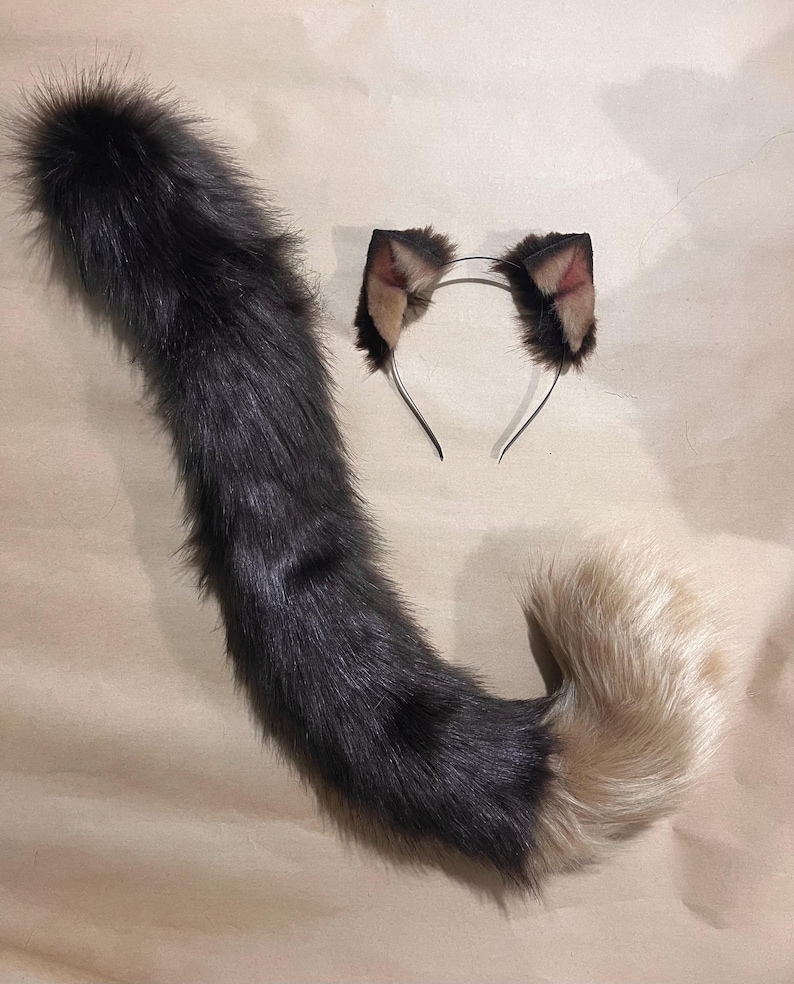 May include: A faux fur cat tail and matching headband with cat ears. The tail is primarily dark gray, transitioning to a light tan at the tip. The ears are dark brown with tan and pink accents, attached to a silver headband.