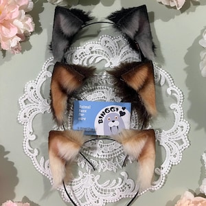 May include: Three faux fur animal ear headbands in gray, brown, and tan, each with a black headband. The ears are displayed on a white lace doily, with a small card that reads "Animal faux fur ears" and the brand name "Binggi".