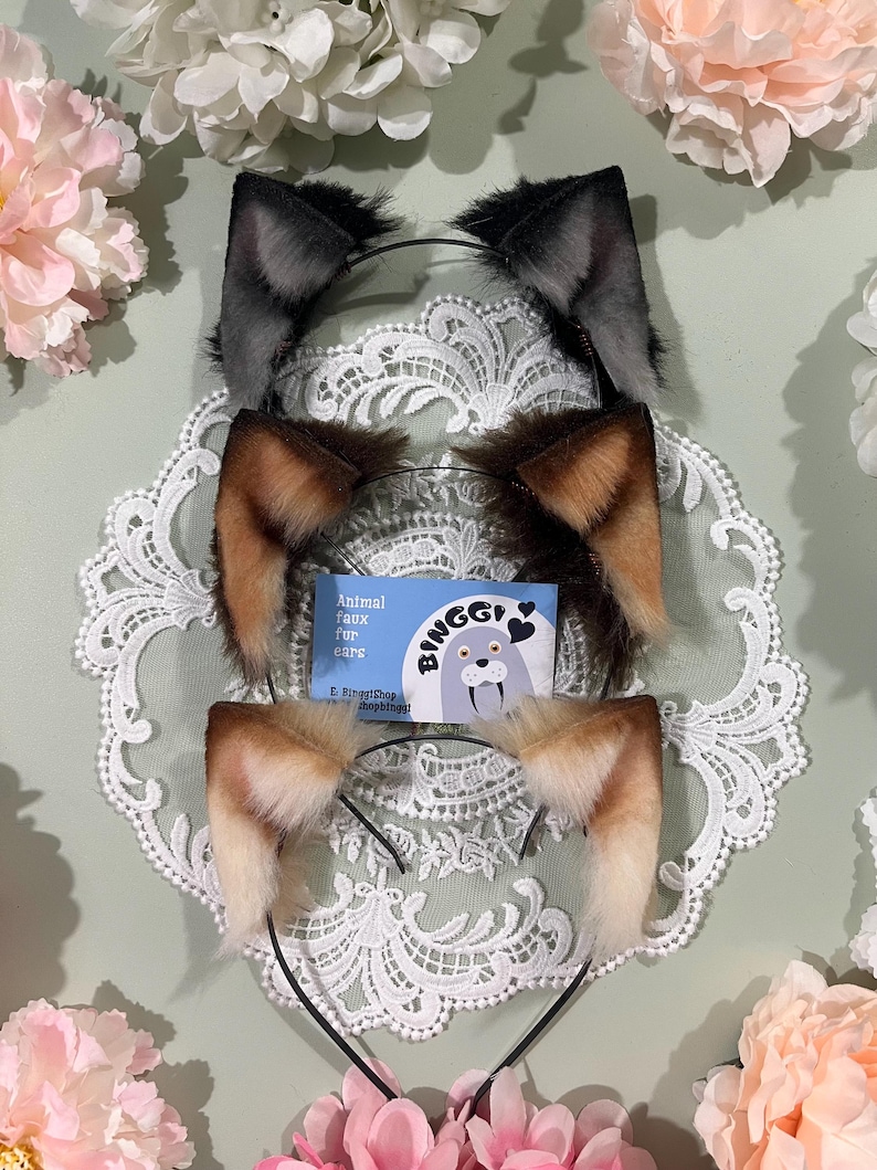 May include: Three faux fur animal ear headbands in gray, brown, and tan, each with a black headband. The ears are displayed on a white lace doily, with a small card that reads "Animal faux fur ears" and the brand name "Binggi".