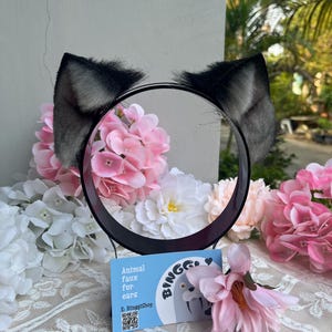 May include: Black and gray faux fur animal ears attached to a black headband. The ears are fluffy and stand upright. A small blue card with the text "Animal faux fur for ears" is in front of the headband. The background includes pink and white flowers.