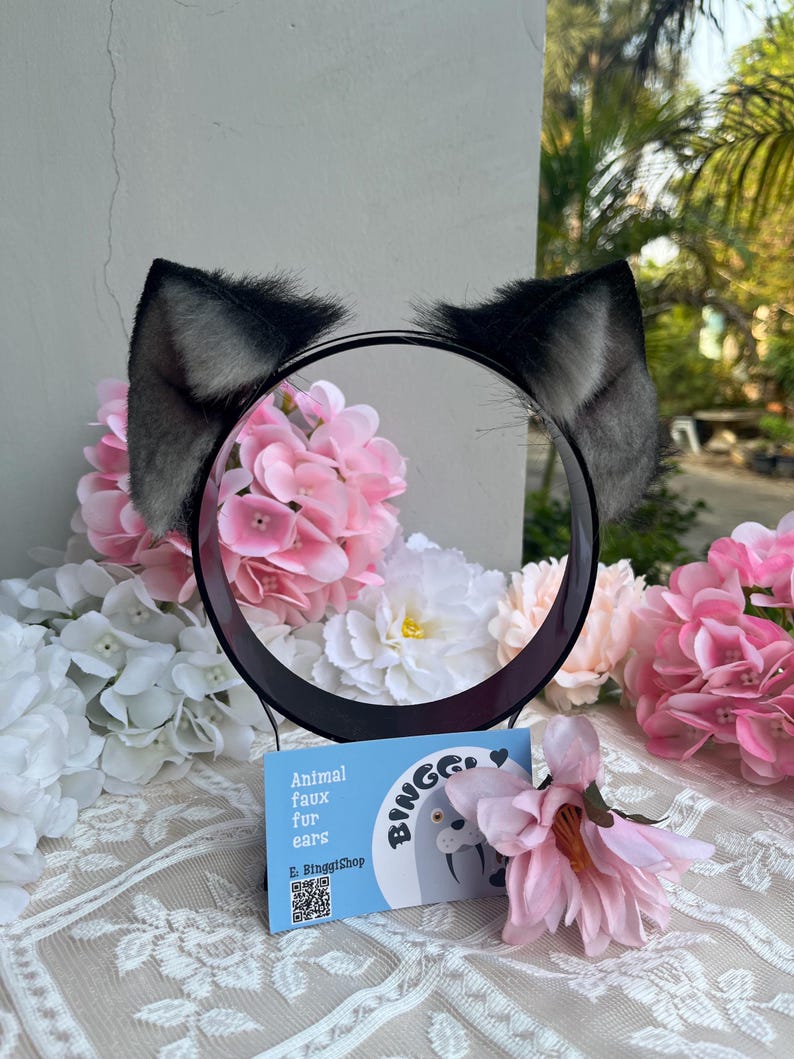 May include: Black and gray faux fur animal ears attached to a black headband. The ears are fluffy and stand upright. A small blue card with the text "Animal faux fur for ears" is in front of the headband. The background includes pink and white flowers.