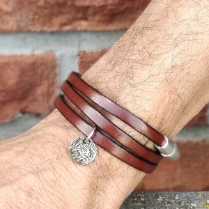 Puede incluir: Un brazalete de cuero marrón con un dije de plata con diseño de huella digital.