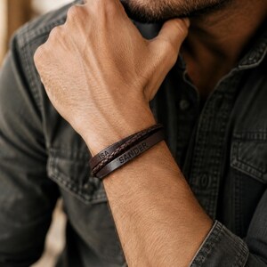 Puede incluir: Una pulsera de cuero marr&oacute;n de varias hebras que presenta un cord&oacute;n trenzado y una banda plana lisa. La banda lisa est&aacute; grabada con el nombre SANDER, mientras que la otra secci&oacute;n est&aacute; grabada con el nombre ISA. La pulsera rodea la mu&ntilde;eca.