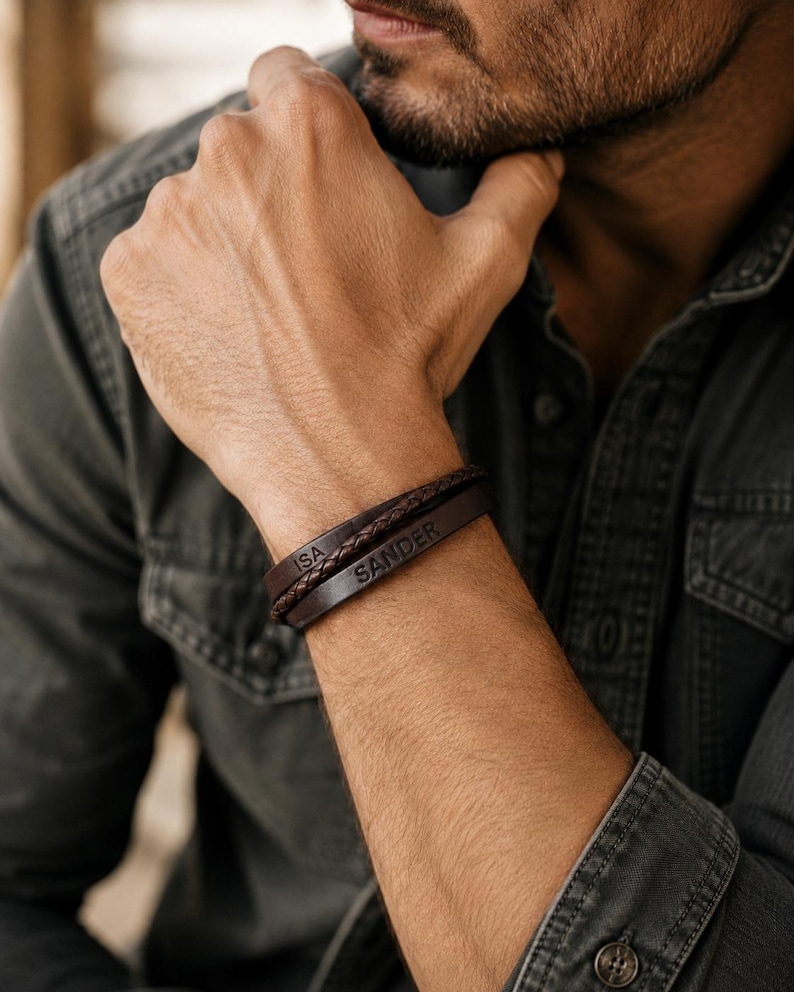 Puede incluir: Una pulsera de cuero marr&oacute;n de varias hebras que presenta un cord&oacute;n trenzado y una banda plana lisa. La banda lisa est&aacute; grabada con el nombre SANDER, mientras que la otra secci&oacute;n est&aacute; grabada con el nombre ISA. La pulsera rodea la mu&ntilde;eca.