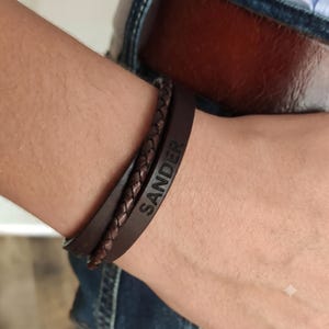 Op de afbeelding: Een donkerbruine leren armband bestaande uit twee verschillende strengen: een gladde platte band met de inscriptie SANDER in hoofdletters en een gevlochten koordstreng. De armband heeft een gelaagd ontwerp met een donkere afwerking.