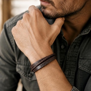 May include: A dark brown leather bracelet featuring two distinct strands: one smooth flat band engraved with the name SANDER and another braided cord strand engraved with the name ISA. The bracelet is worn on a wrist, showcasing the dark brown leather.