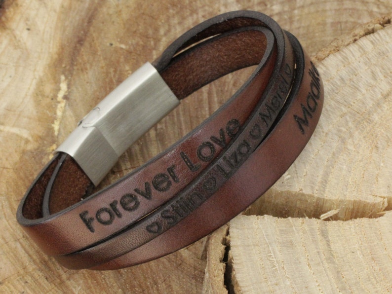 K&ouml;nnte beinhalten: Braunes Lederarmband mit einem silbernen Verschluss. Das Armband ist mit den Worten "Forever Love" und "Still Like a Melody" graviert.