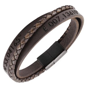 Puede incluir: Una pulsera de cuero marr&oacute;n oscuro que presenta una tira de cuero trenzado y una tira de cuero lisa grabada con las coordenadas N 53.04165&deg; E 007.13610&deg;. La pulsera se asegura con un cierre magn&eacute;tico rectangular de metal negro.