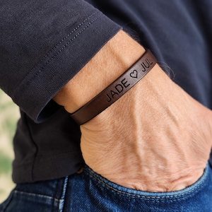 Puede incluir: Una pulsera de cuero marrón oscuro con el texto grabado JADE seguido de un símbolo de corazón y el nombre JUL. La banda es lisa y rodea la muñeca, mostrando letras oscuras y claras sobre la superficie del cuero.