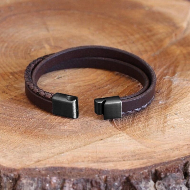 Puede incluir: Una pulsera de cuero marr&oacute;n oscuro con un dise&ntilde;o de varias hebras, que incluye una banda de cuero liso y una banda de cuero trenzado. La pulsera cuenta con un cierre rectangular de metal negro mate que se muestra en posici&oacute;n abierta.
