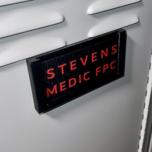 Könnte beinhalten: Ein schwarzes Namensschild mit dem Text "STEVENS MEDIC FPC" in roter Schrift. Das Namensschild ist auf einer hellgrauen Oberfläche mit einer Reihe horizontaler, weißer, rechteckiger Lüftungsschlitze angebracht.