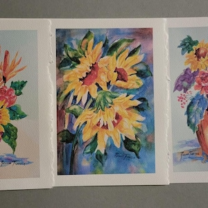 May include: Three watercolor paintings of floral arrangements in vases. The first painting features a pink vase with a bouquet of yellow, orange, red, and blue flowers. The second painting features a blue vase with a bouquet of yellow sunflowers. The third painting features an orange vase with a bouquet of yellow sunflowers, red and purple flowers, and green leaves.