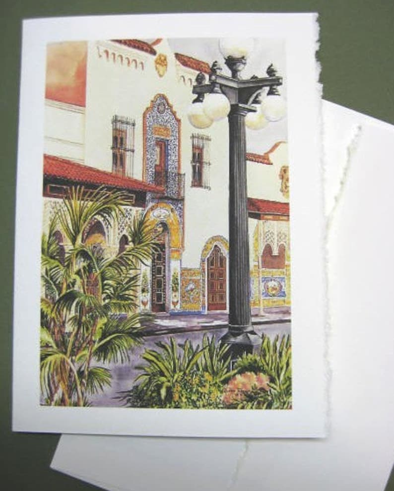 Op de afbeelding: Een aquarel van een gebouw met een zwarte lantaarnpaal ervoor. Het gebouw is geschilderd in tinten wit, geel en blauw. De lantaarnpaal heeft een zwarte paal en een witte bol. De aquarel is geschilderd tegen een achtergrond van een blauwe lucht en groene bomen.