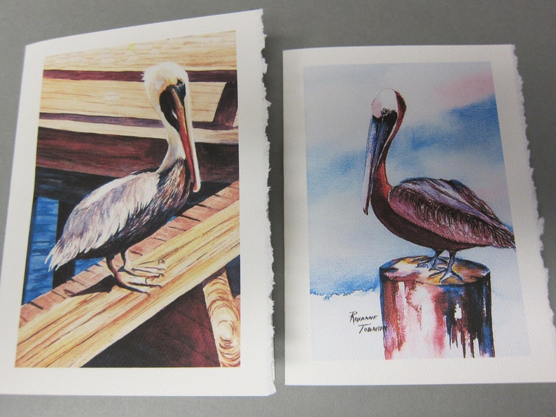 May include: Two watercolor paintings of brown pelicans. The pelican on the left is perched on a wooden dock with blue water visible in the background. The pelican on the right is perched on a red and blue wooden post against a blue and white sky background. The artist's signature is visible in the bottom right corner of the painting on the right.