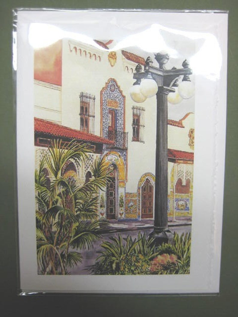 Op de afbeelding: Een aquarel van een wit gebouw met een betegelde gevel en een lantaarnpaal ervoor. Het gebouw heeft een rood pannendak en een balkon met een smeedijzeren balustrade. De lantaarnpaal is zwart met een gouden top en heeft meerdere lampen. Op de voorgrond staan groene planten.