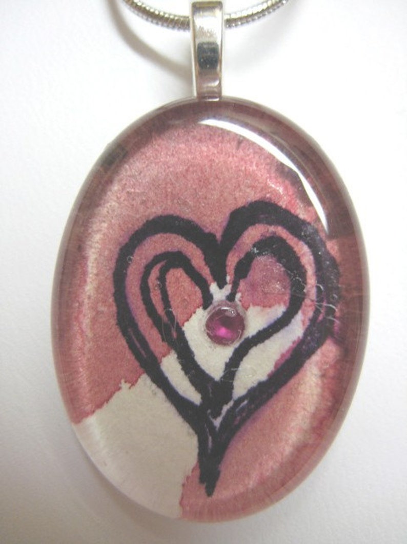 May include: A pink glass pendant with a black heart design and a small pink gem in the center. The pendant is attached to a silver chain.