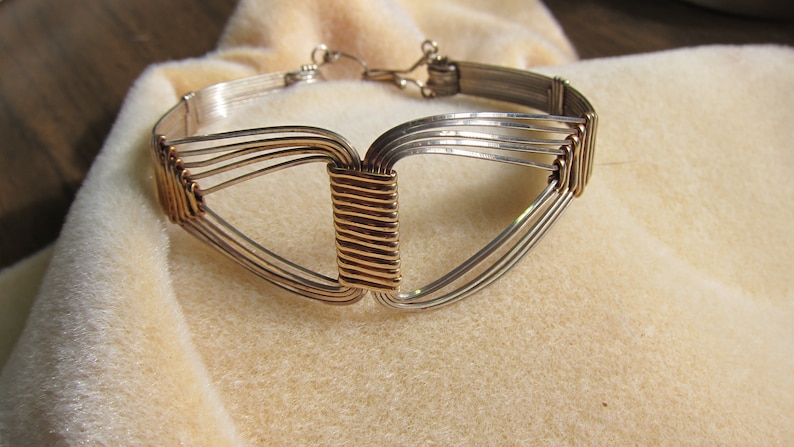 May include: A silver and gold wire bracelet with a geometric design. The bracelet is made of multiple strands of wire that are woven together to create a triangular shape. The center of the bracelet is a gold wire that is woven into a rectangular shape.