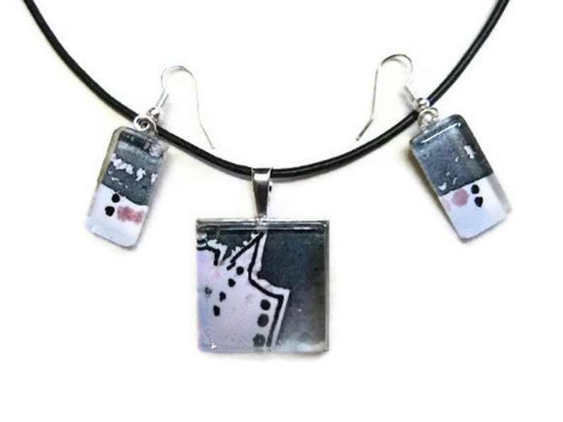 May include: A black necklace with a silver pendant and matching earrings. The pendant and earrings are rectangular and feature a white and gray abstract design with pink accents.