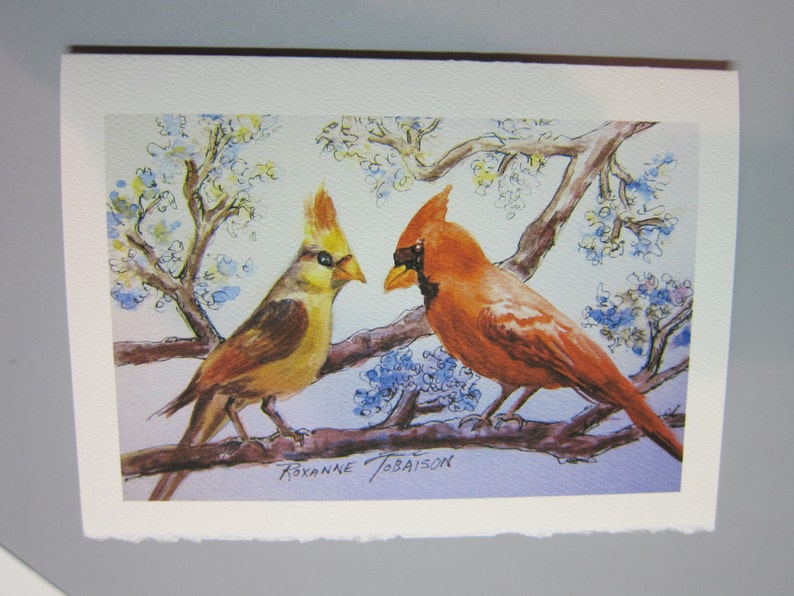 K&ouml;nnte beinhalten: Ein Aquarellbild von zwei Kardin&auml;len, die auf einem Ast sitzen. Der m&auml;nnliche Kardinal ist rot und der weibliche Kardinal ist gelb. Der Hintergrund ist hellblau mit wei&szlig;en Bl&uuml;ten.