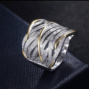 Puede incluir: Un anillo plateado con un diseño tejido, acentuado con bandas doradas. El anillo está incrustado con pequeñas piedras preciosas claras, creando un efecto brillante. El anillo está sobre un fondo azul oscuro.