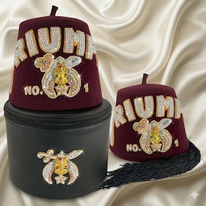 May include: Burgundy fez hats with the word "TRIUMPH" in rhinestone letters. The hats feature an ornate emblem with the number "1" and are displayed with a black carrying case. A black tassel hangs from one hat.